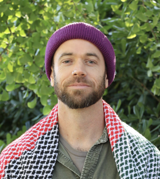 Portrait photo — bearded man in purple beanie and patterned shawl, leafy background